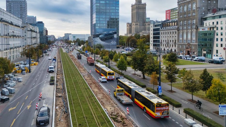 Ruchliwe skrzyżowanie z autobusami i samochodami, w tle nowoczesne budynki i wieżowiec z zegarem.