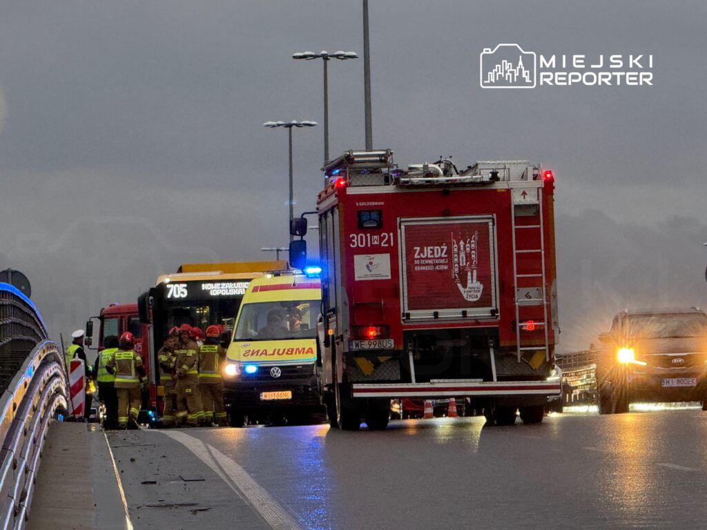 Grupa strażaków w ubraniach ochronnych rozmawia obok karetki i wozu strażackiego na deszczowej drodze.