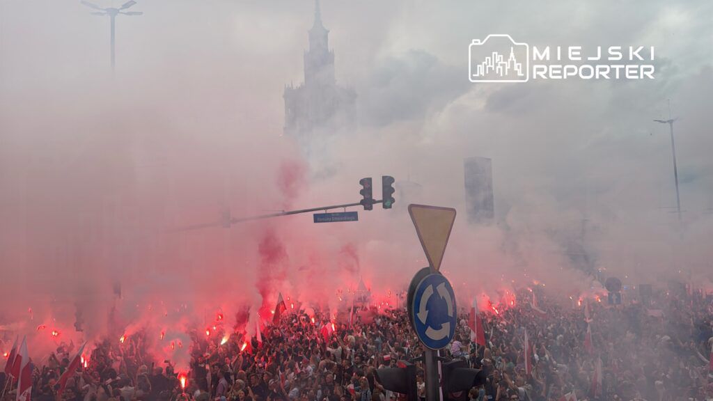 Tłum ludzi trzymających flagi, otoczony czerwonymi racami, w mglistym miejskim otoczeniu.