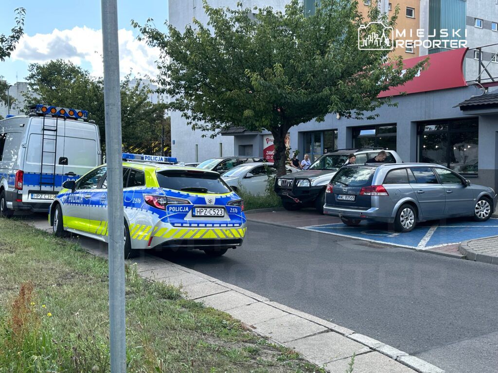 Radiowóz policyjny i furgonetka z niebieskimi światłami stoją na parkingu obok budynku, obok kilku zaparkowanych samochodów.