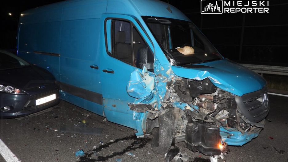 Zniszczony niebieski furgon z uszkodzoną przednią częścią stojący na drodze, obok widać uszkodzony samochód osobowy.