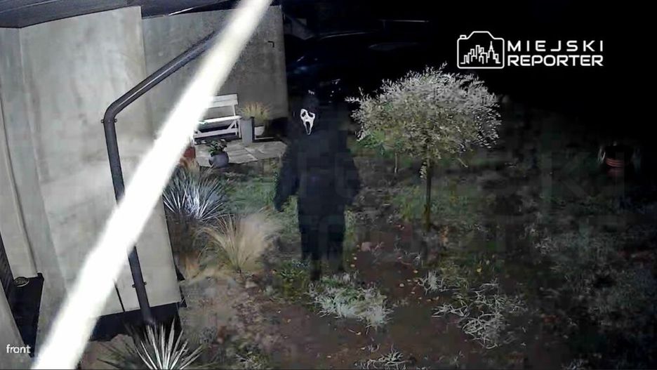 Osoba w czarnym stroju z maską stoi na podjeździe, otoczona przez roślinność w nocy.