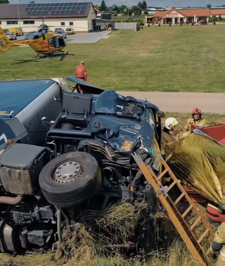 Pożarnicy pracują przy przewróconym ciężarówce, a helikopter lądował w tle na trawiastej przestrzeni.