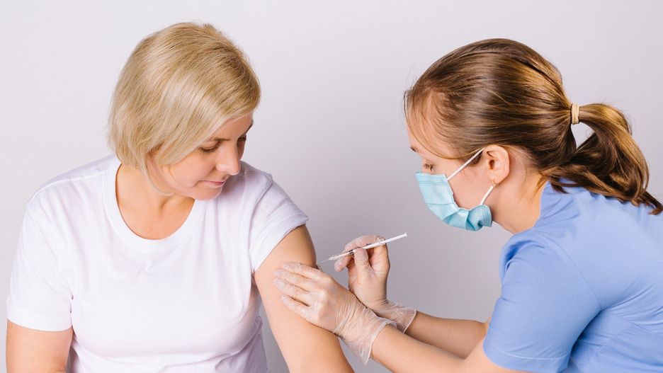 Vaccination of the population. Young nurse puts covid vaccine on 19 senior blonde woman against white background.