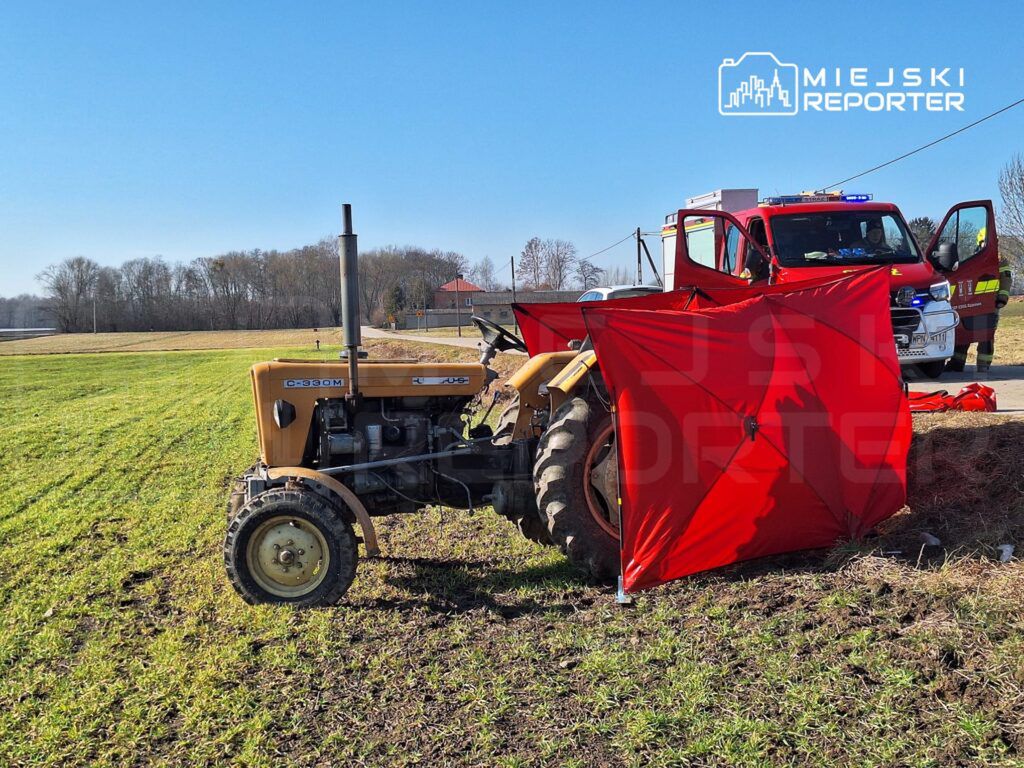 Traktor na polu z czerwoną osłoną, obok stoi wóz strażacki z otwartymi drzwiami.