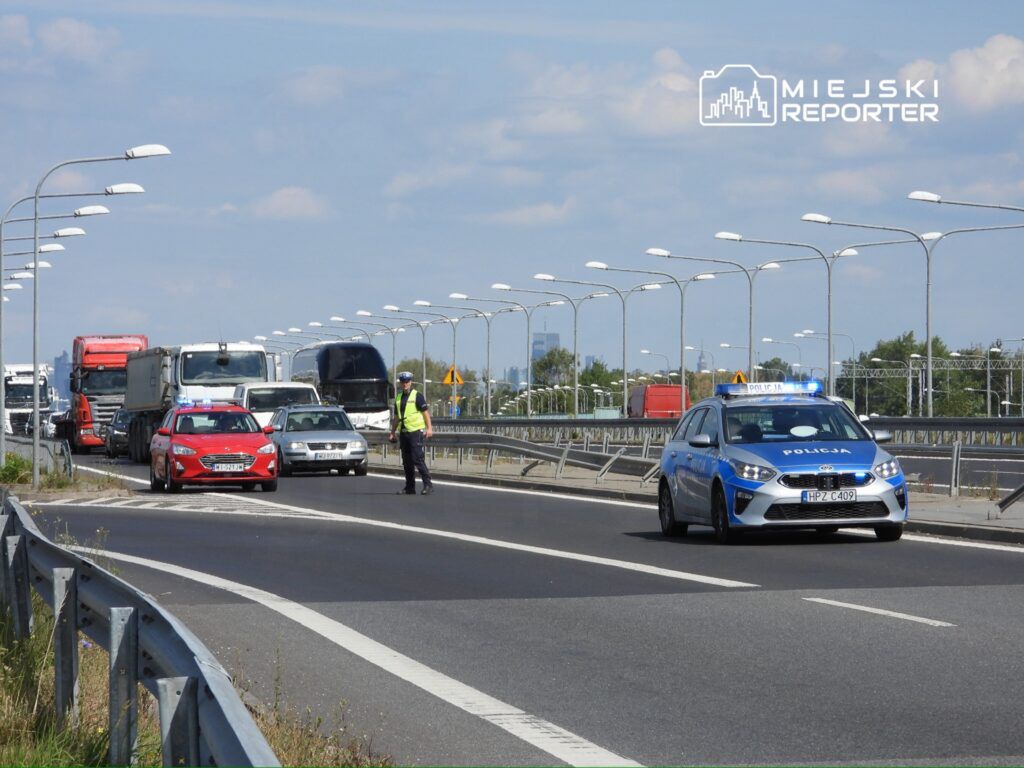 Policjant w kamizelce odblaskowej kieruje ruchem na drodze, obok zaparkowane pojazdy, w tym radiowóz z włączonymi sygnałami.