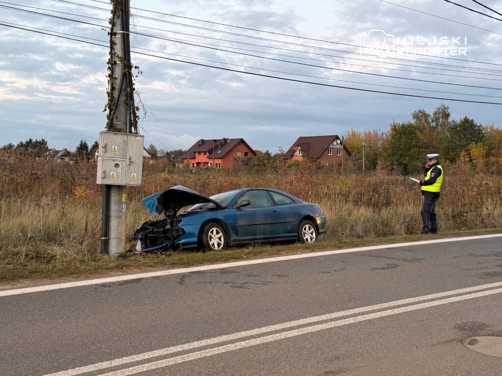Nieoznakowany samochód osobowy z uszkodzoną maską stoi przy słupie energetycznym, obok policjant sporządzający notatki.