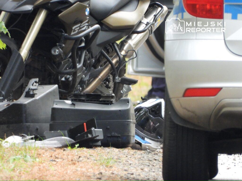 Motocykl leży na poboczu drogi, obok niego znajduje się kask oraz policyjny samochód.