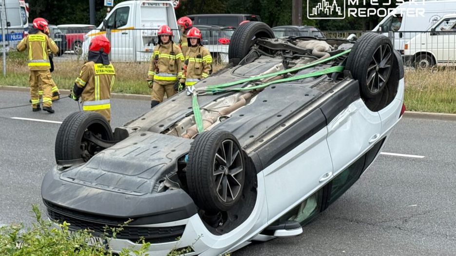 Kierowca w samochodzie osobowym leżącym na dachu, obok strażacy w mundurach pracujący przy akcji ratunkowej na drodze.