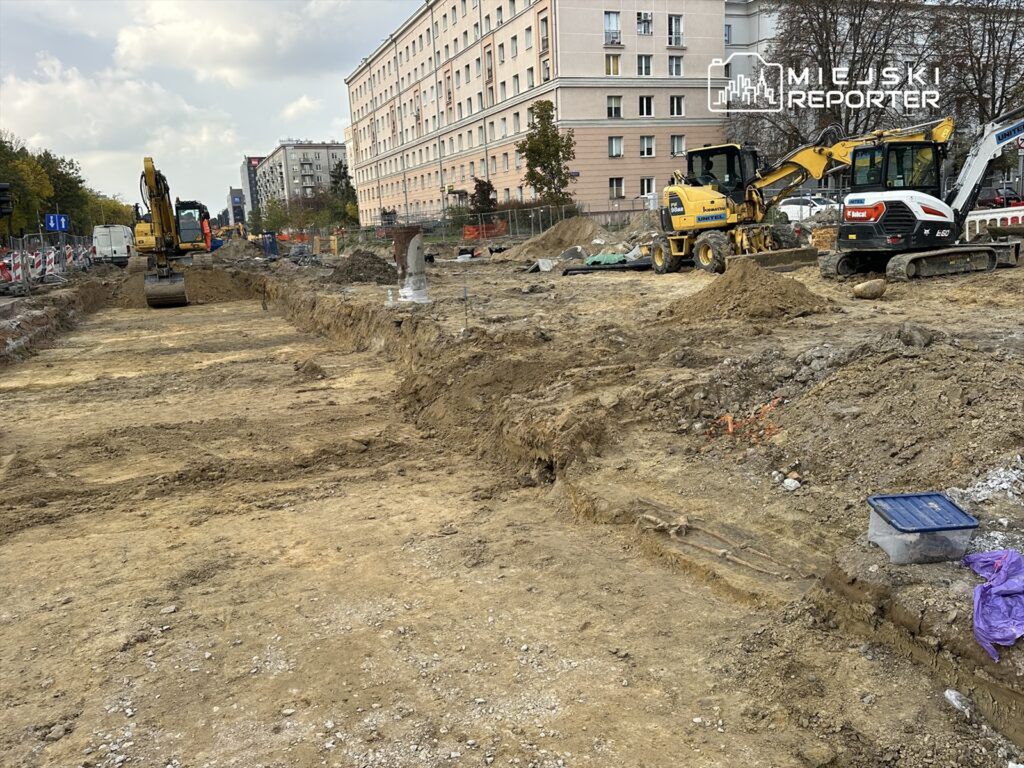 Wykopy budowlane z ciężkim sprzętem na placu budowy w miejskim otoczeniu, w tle bloki mieszkalne.