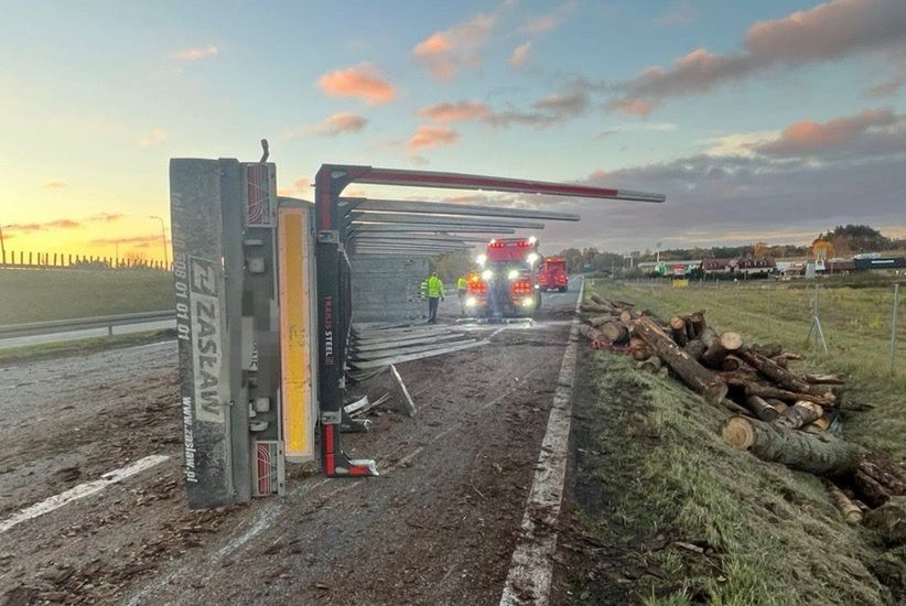 Przewrócony kontener na drodze, obok pracujący strażacy i leżące na poboczu kłody drewna.