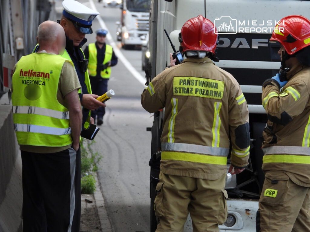 Kierowca w kamizelce odblaskowej rozmawia z policjantem, obok strażacy kontrolują uszkodzony pojazd na drodze.