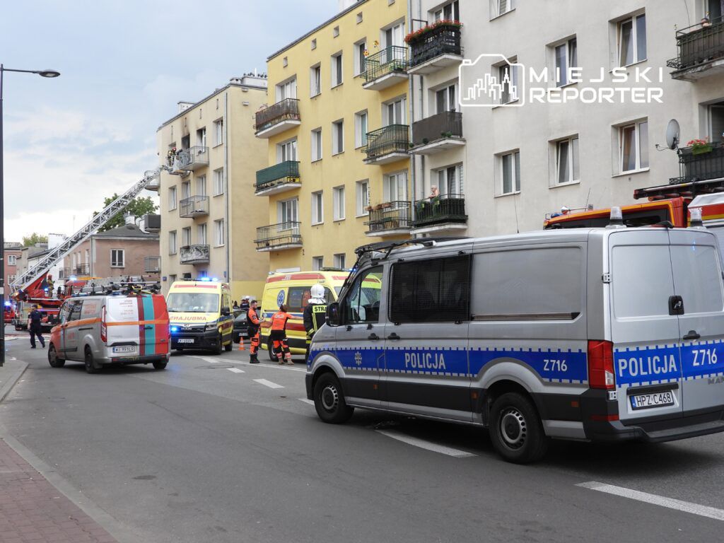Na ulicy z widocznymi pojazdami straży pożarnej i ambulansami, ratownicy interweniują przy budynku mieszkalnym.