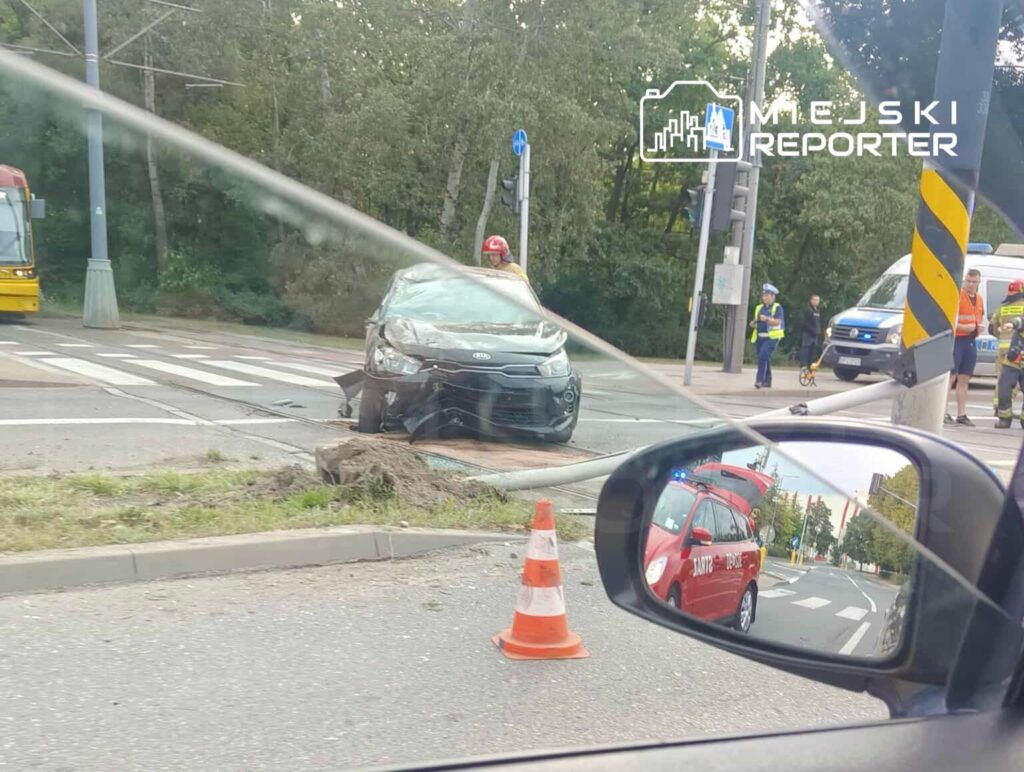 Samochód osobowy z uszkodzoną przednią częścią stoi na chodniku, obok grupy ratowników i policjantów.