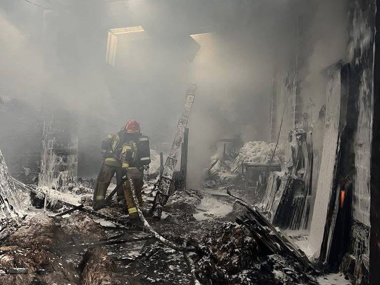 Strażacy w hełmach gaszą pożar w zadymionym pomieszczeniu, otoczonym przez zniszczone materiały.