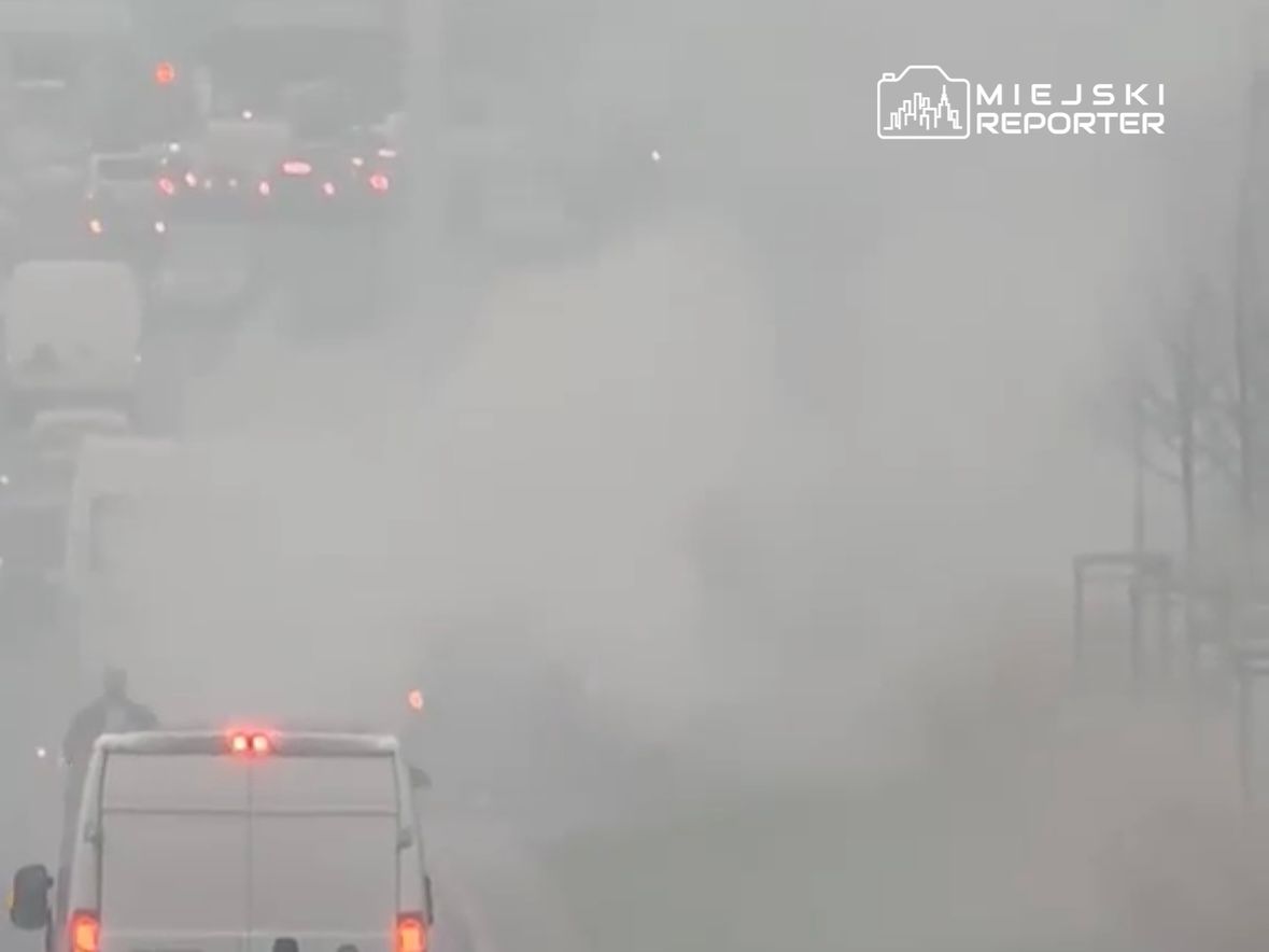 Gęsty smog ogranicza widoczność na ruchliwej drodze, gdzie stoją samochody i dostawczaki.