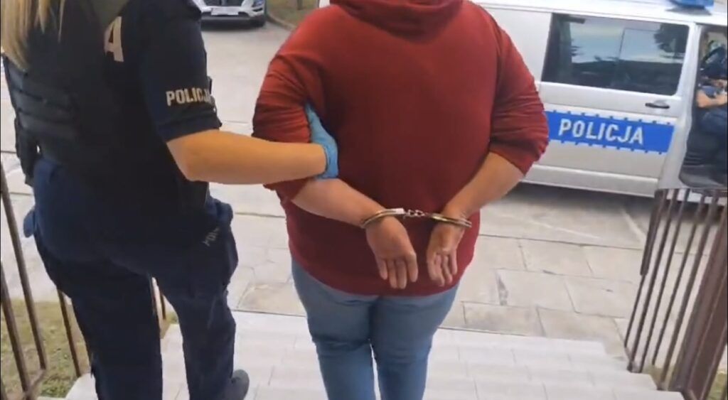 Kobieta w czerwonym swetrze jest prowadzona przez policjantkę w mundurze, ręce ma skute w kajdankach.