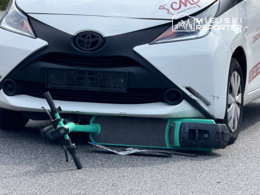 Zielona hulajnoga elektryczna leży pod przednim zderzakiem białego samochodu osobowego.