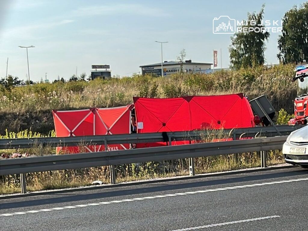 Czerwony namiot ratunkowy ustawiony obok drogi, w tle widoczne pojazdy i zieleń przy autostradzie.
