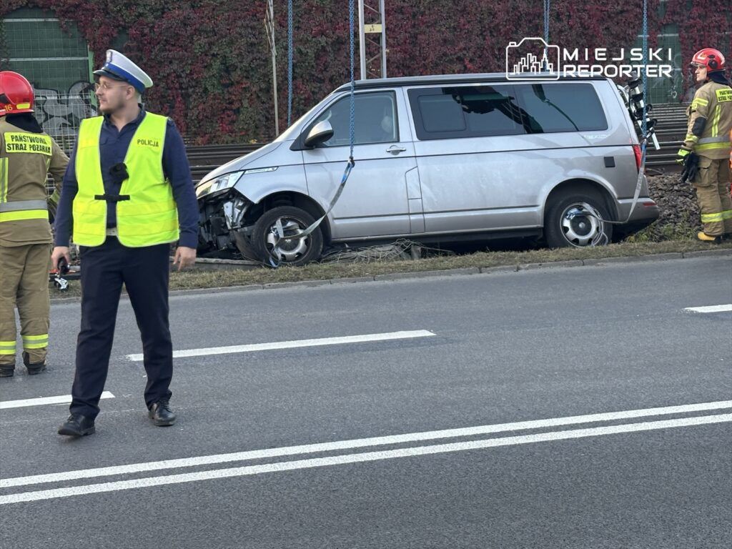 Mężczyzna w mundurze policyjnym obserwuje zdarzenie drogowe obok uszkodzonego vana, na którym pracują strażacy.