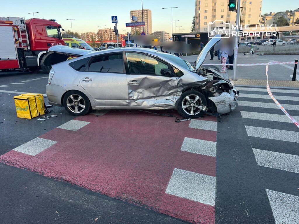 Zniszczony samochód osobowy zaparkowany na przejściu dla pieszych, obok torów, z widocznymi torbami dostawczymi.