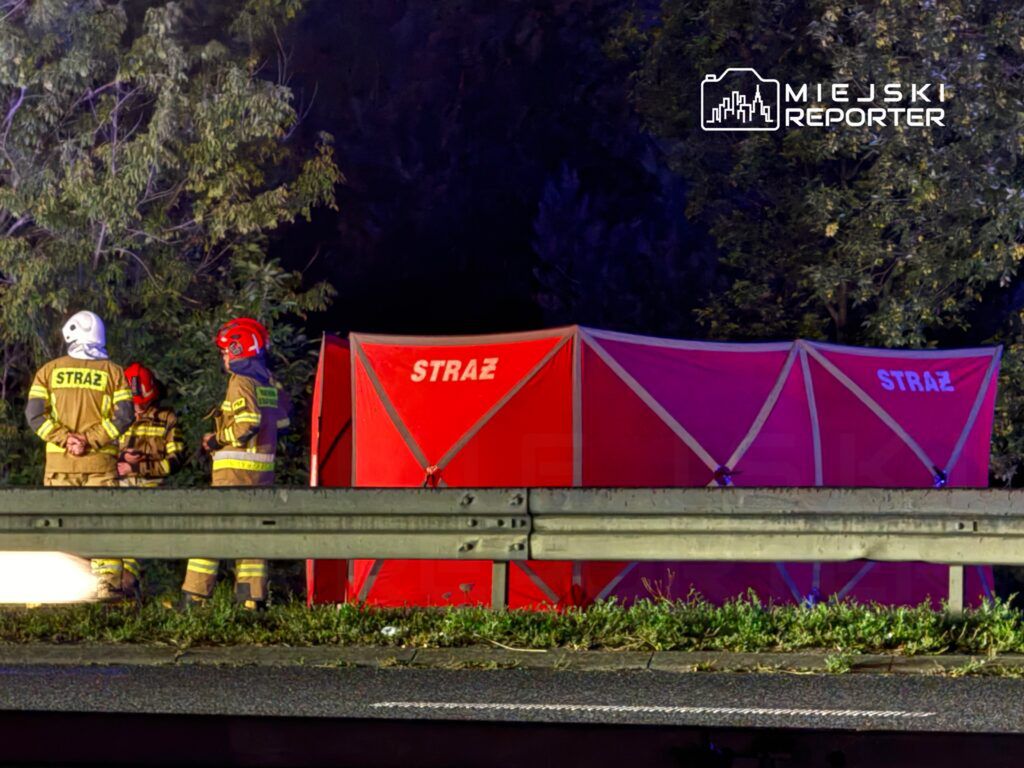 Grupa strażaków w mundurach rozmawia obok czerwonego namiotu z napisem "STRAŻ" przy drodze.
