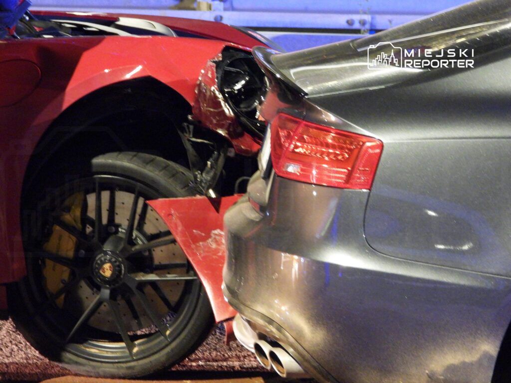 Czerwony samochód sportowy z uszkodzoną przednią częścią zderza się z tyłem szarego auta, na tle nocnej ulicy.