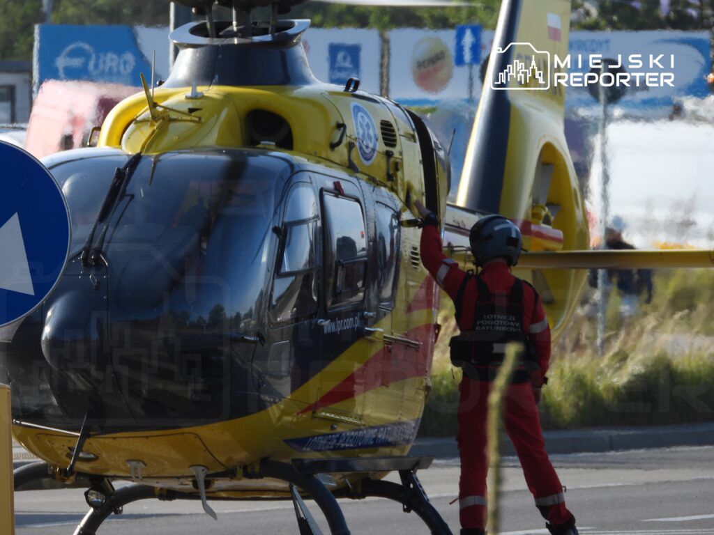 Pilot w czerwonym kombinezonie z logo Lotniczego Pogotowia Ratunkowego otwiera drzwi helikoptera na lądowisku.