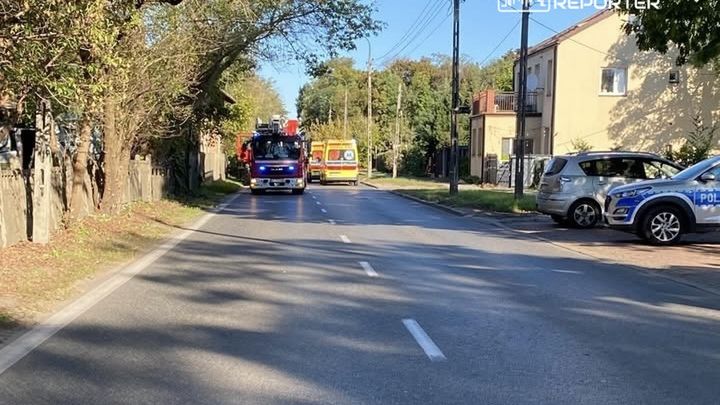 Na drodze widać woz z strażą pożarną oraz ambulans, a obok parkują radiowóz policyjny. Po obu stronach drogi rosną drzewa.