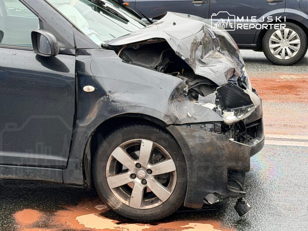 Czarny samochód osobowy z uszkodzoną przednią częścią stoi na drodze, obok widać inne pojazdy.