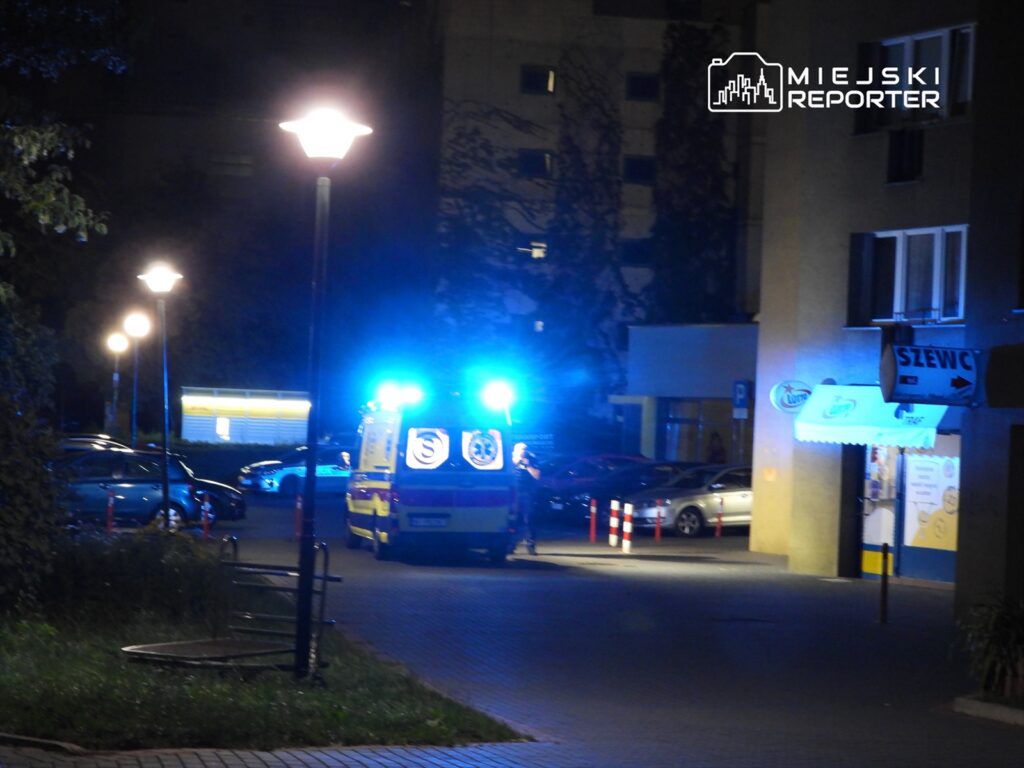 Karetka pogotowia z włączonymi sygnałami świetlnymi zatrzymana na oświetlonym parkingu nocą.