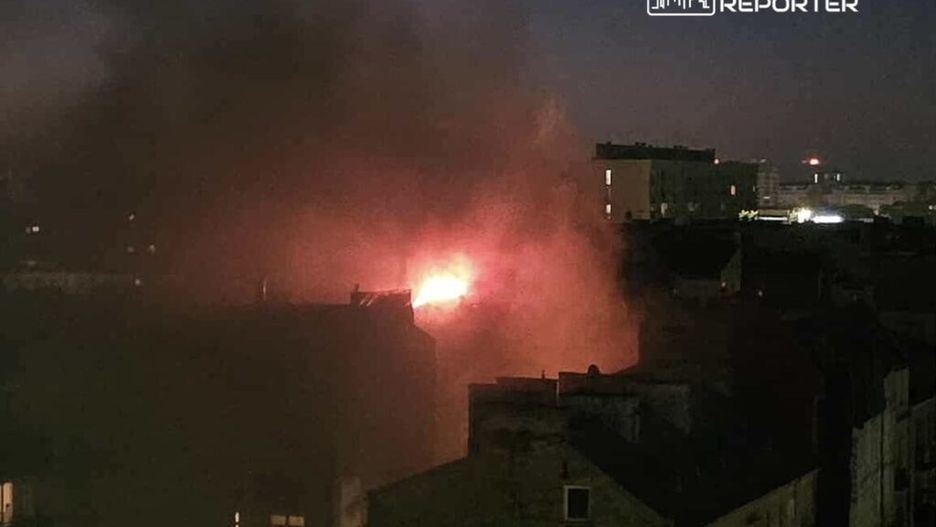 Płomienie i dym unoszą się nad budynkami w nocnej scenerii miejskiej.