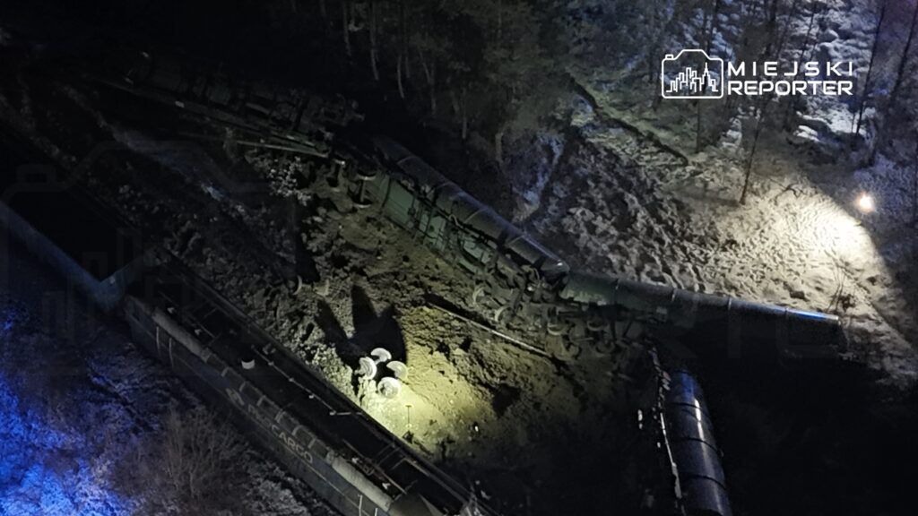 Zniszczony pociąg towarowy leżący na boku w zaśnieżonym terenie, oświetlony przez latarki.