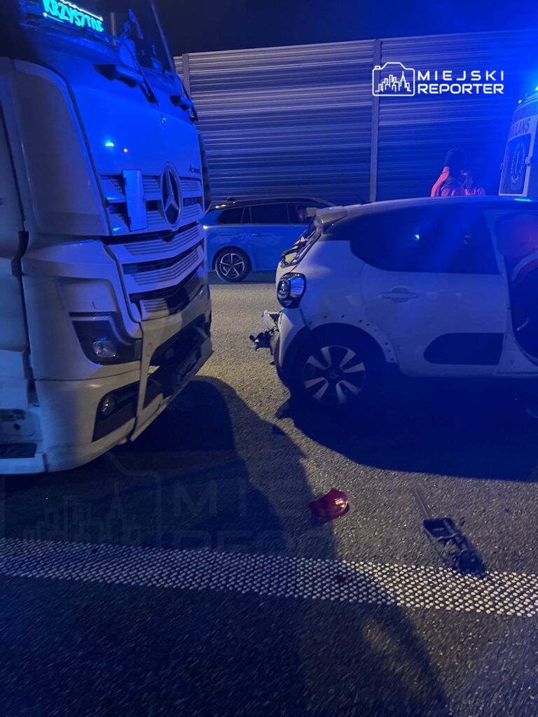 W nocy na drodze stoi ciężarówka obok uszkodzonego samochodu osobowego, a na jezdni leżą fragmenty lampy.