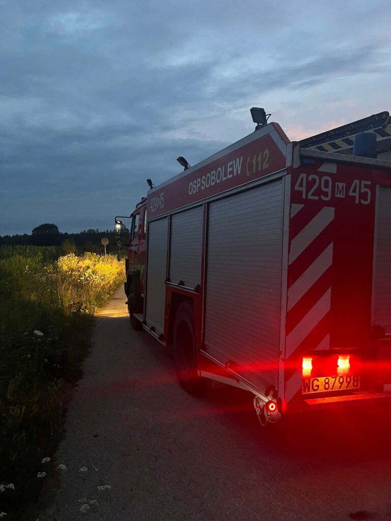 Czerwony wóz strażacki OSP Sobolew zaparkowany na wąskiej drodze, oświetlający otoczenie w świetle zachodzącego słońca.