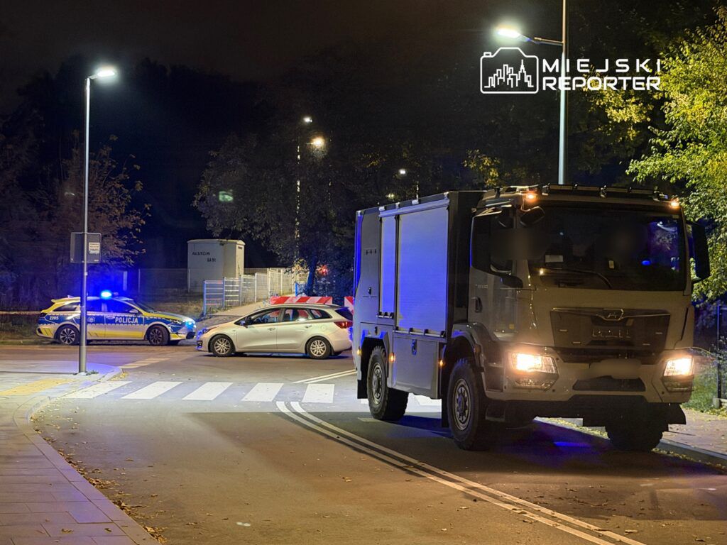 Policyjny radiowóz z włączonymi sygnałami świetlnymi oraz duży pojazd służbowy na oświetlonej ulicy nocą.