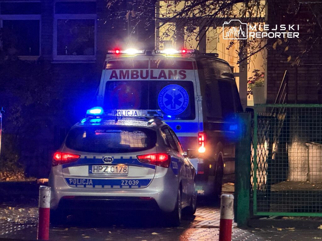 Radiowóz policji i ambulans z włączonymi sygnałami świetlnymi stoją przed budynkiem mieszkalnym w nocy.