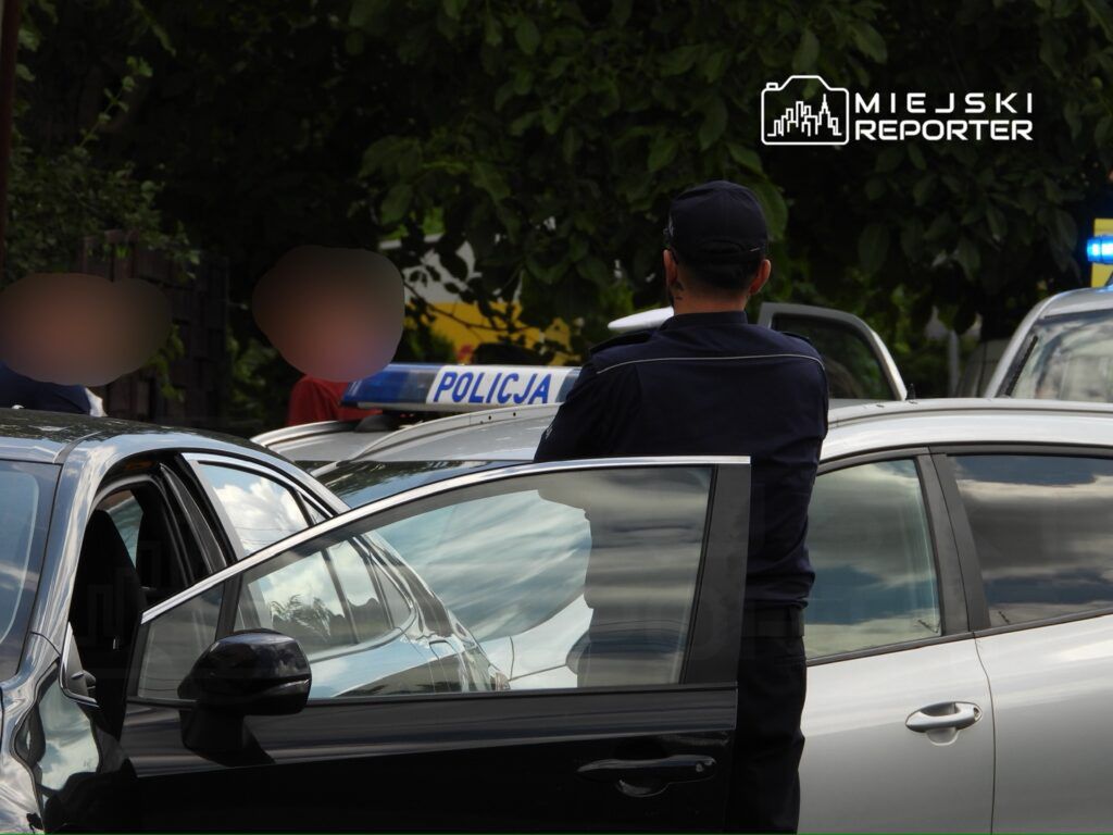 Policjant rozmawia z osobami stojącymi obok zaparkowanych samochodów w miejskim otoczeniu.