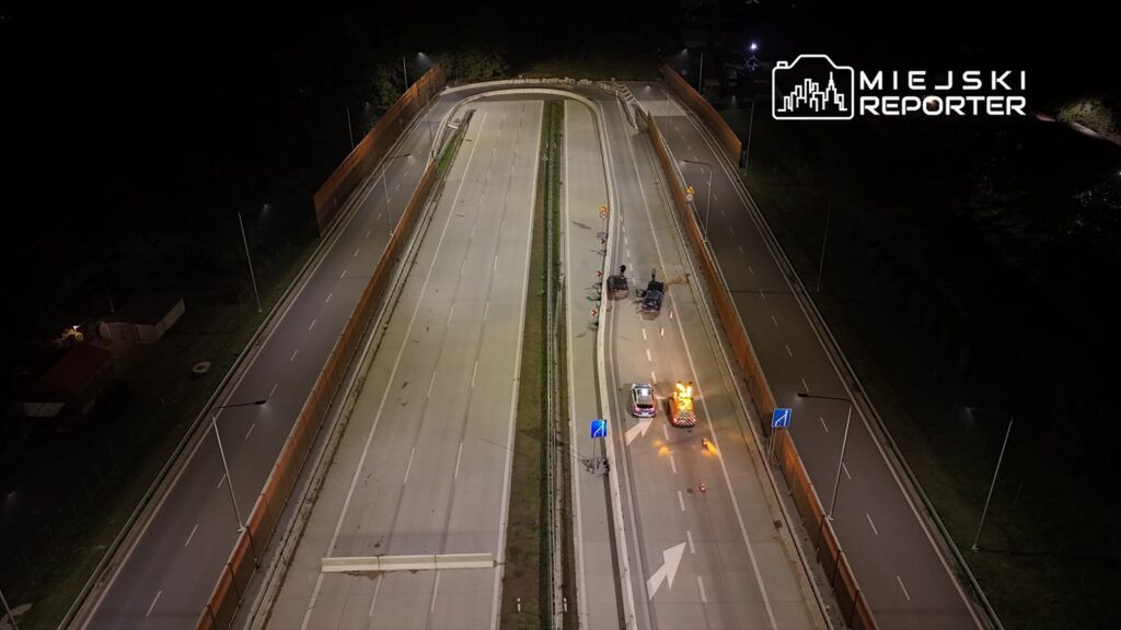 Na drodze nocą stoją dwa samochody służbowe, a w tle widoczne są bariery i lampy oświetlające jezdnię.