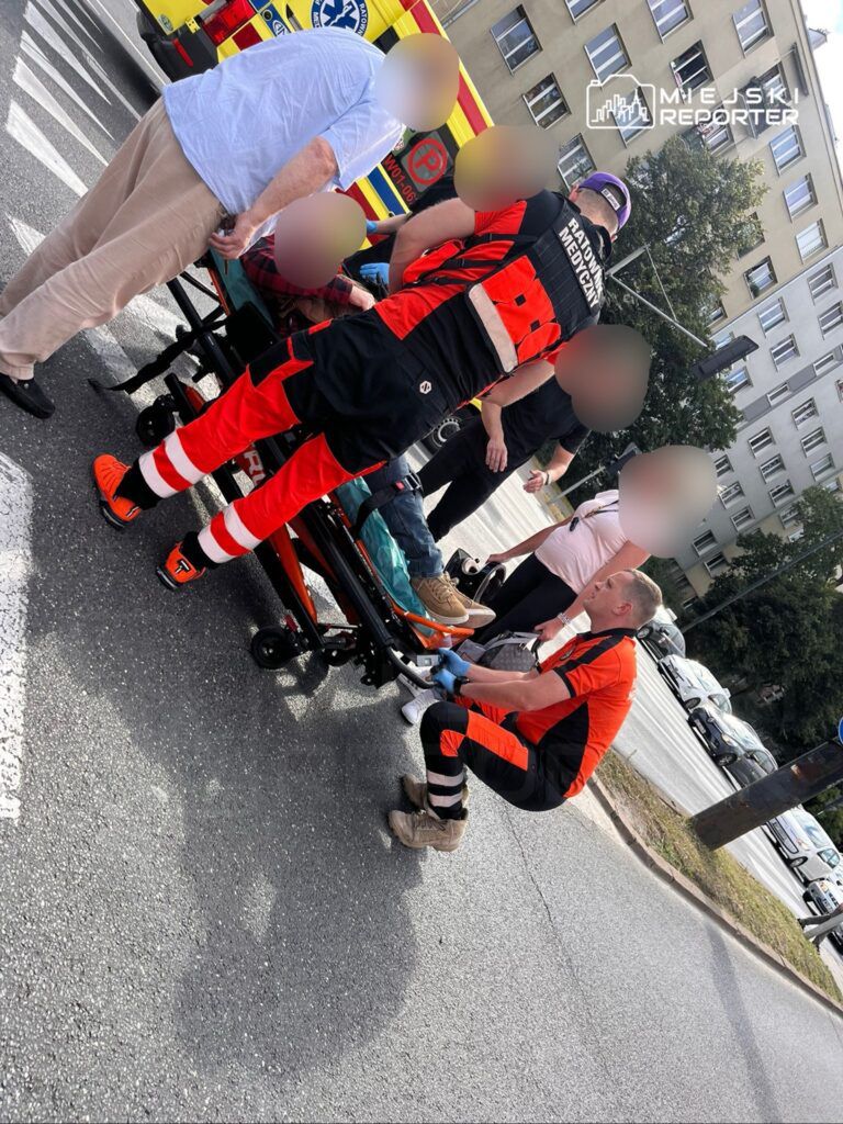 Na zdjęciu widać interwencję medyczną na ulicy. Ratownicy medyczni udzielają pomocy osobie leżącej na noszach, podczas gdy świadkowie obserwują sytuację. W tle widoczna karetka pogotowia oraz ruch uliczny. Akcja ma miejsce w mieście, w słoneczny dzień.