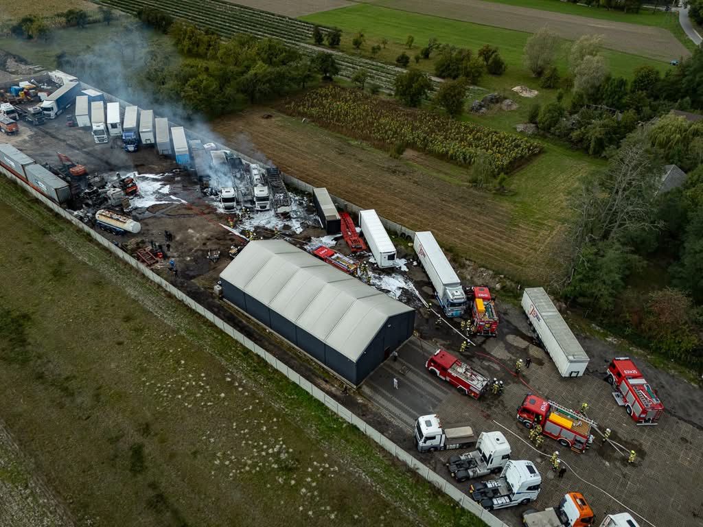 Pożar w magazynie, w którym widać strażaków gaszących ogień oraz uszkodzone ciężarówki. W tle pola i zieleń.