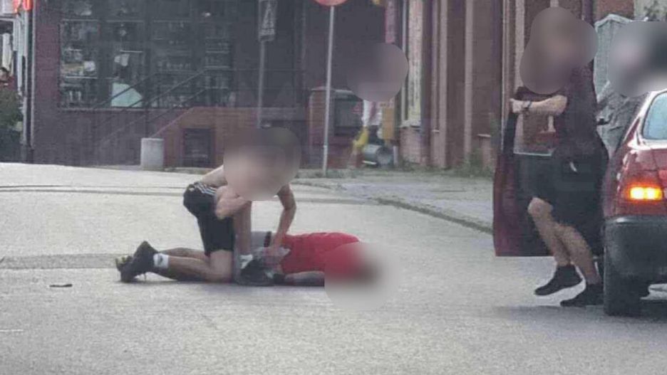 Dwaj mężczyźni na ulicy, jeden leży na ziemi w czerwonym ubraniu, a drugi znajduje się nad nim. W tle widoczny jest sklep i znaki drogowe.