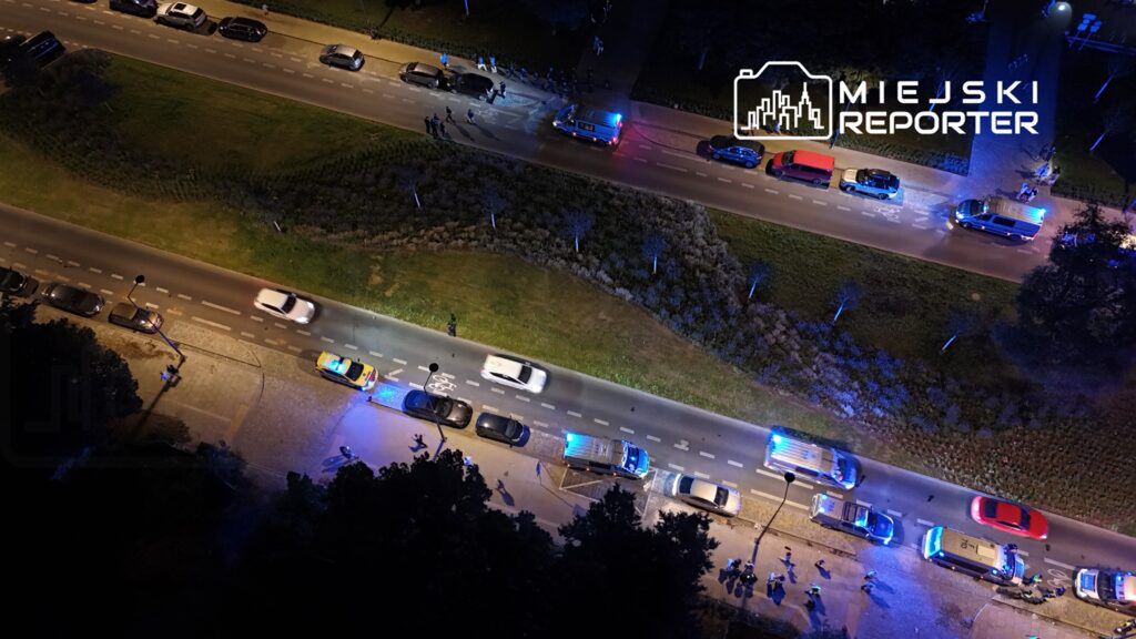 Grupa policjantów interweniuje na oświetlonej ulicy, obok zaparkowanych samochodów, z włączonymi sygnałami świetlnymi.