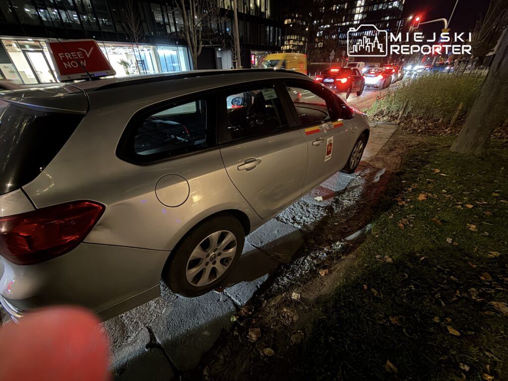 Srebrny samochód zaparkowany na chodniku, obok widoczny znak "FREE NOW" oraz ruchliwa ulica w tle.