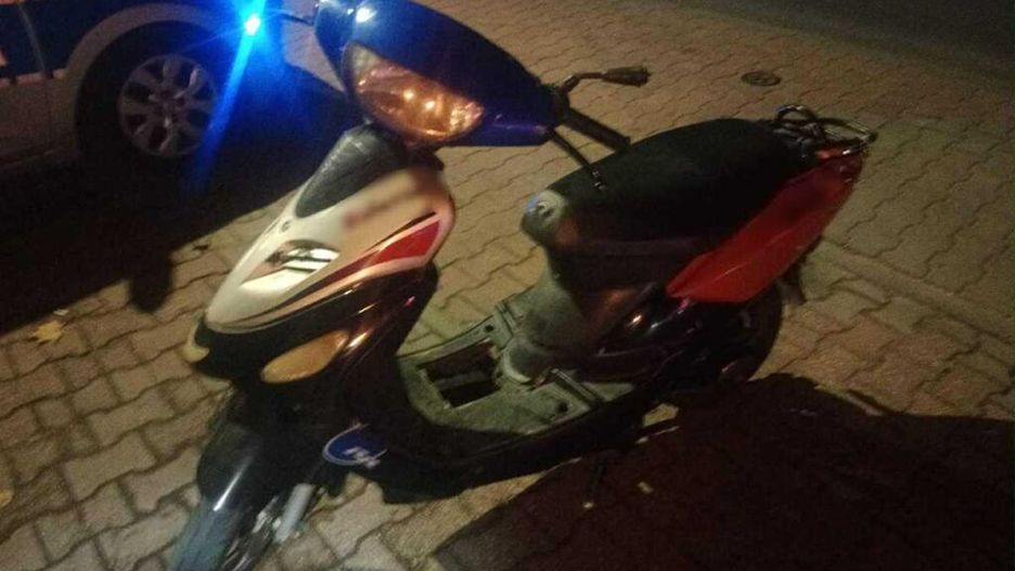 Skuter zaparkowany na kostkowanej nawierzchni obok radiowozu policyjnego z włączonymi światłami.