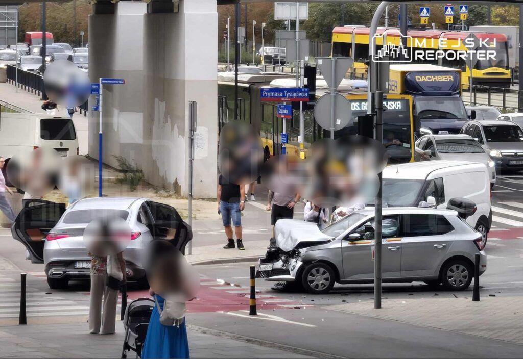 Zderzenie dwóch samochodów na ruchliwej ulicy, obok grupy osób obserwujących zdarzenie. W tle widoczne przystanki komunikacji miejskiej.