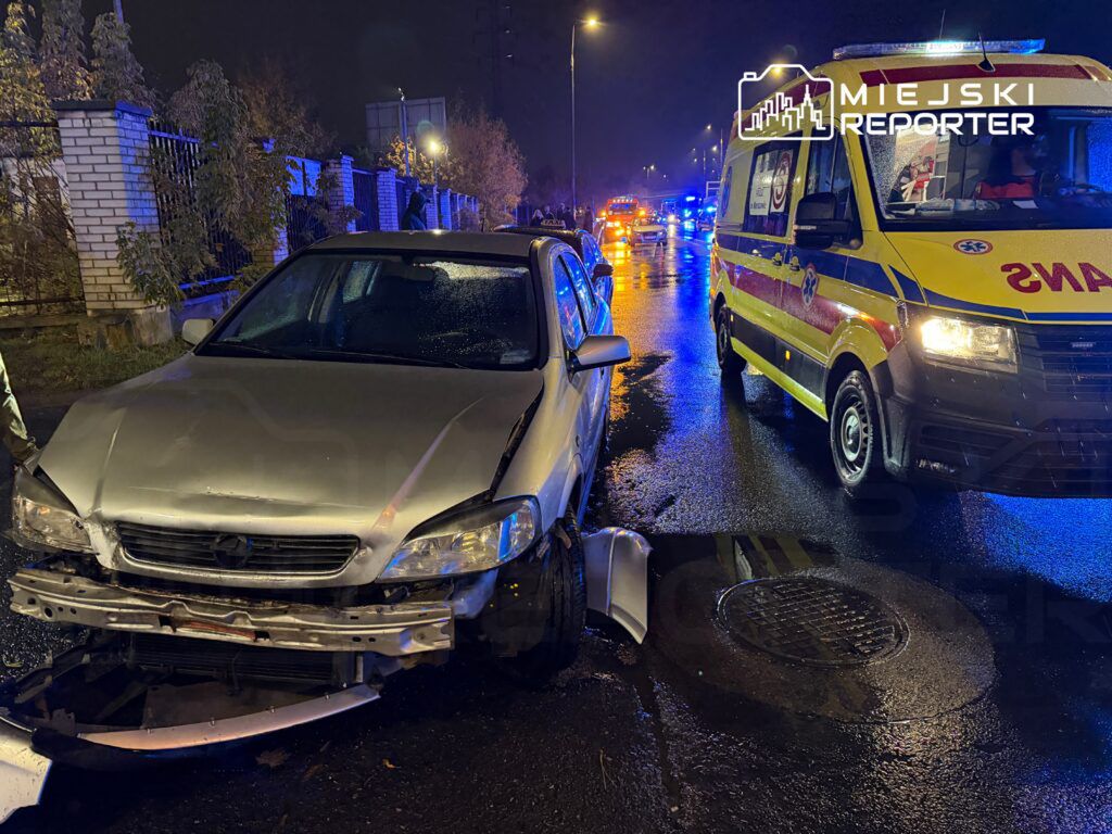 Rozbity samochód osobowy z uszkodzoną przednią częścią stoi na mokrej ulicy, obok niego widać karetkę pogotowia.