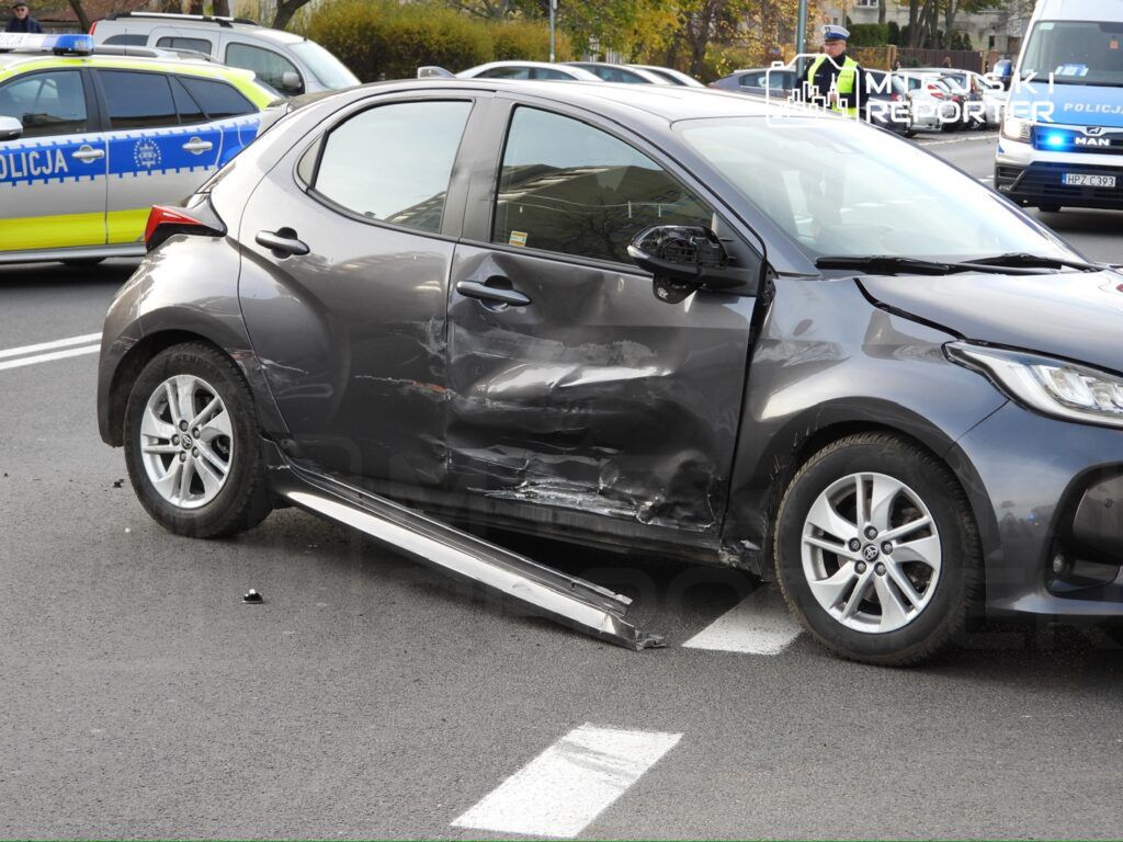 Czarny samochód osobowy z uszkodzeniami bocznymi stoi na drodze, obok widać policyjny radiowóz.