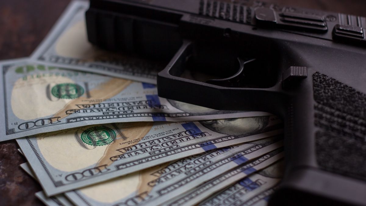 Black gun on American dollars background. Military industry, war, global arms trade, weapon sale, contract killing and crime conceptBlack gun on American dollars background. Military industry, war, global arms trade, weapon sale, contract killing and crime concept.Ildar Imashev