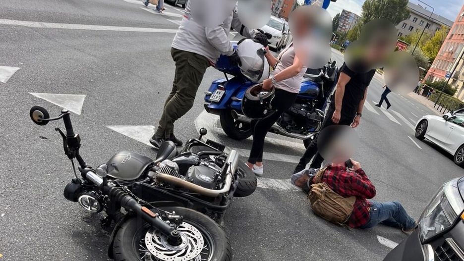 Na zdjęciu widoczny jest wypadek drogowy, w którym przewrócony motocykl leży na jezdni. Obok motocykla stoją osoby, które udzielają pomocy poszkodowanemu leżącemu na ziemi. W tle widać inne pojazdy oraz przechodniów. Zdarzenie miało miejsce w mieście, w godzinach popołudniowych.